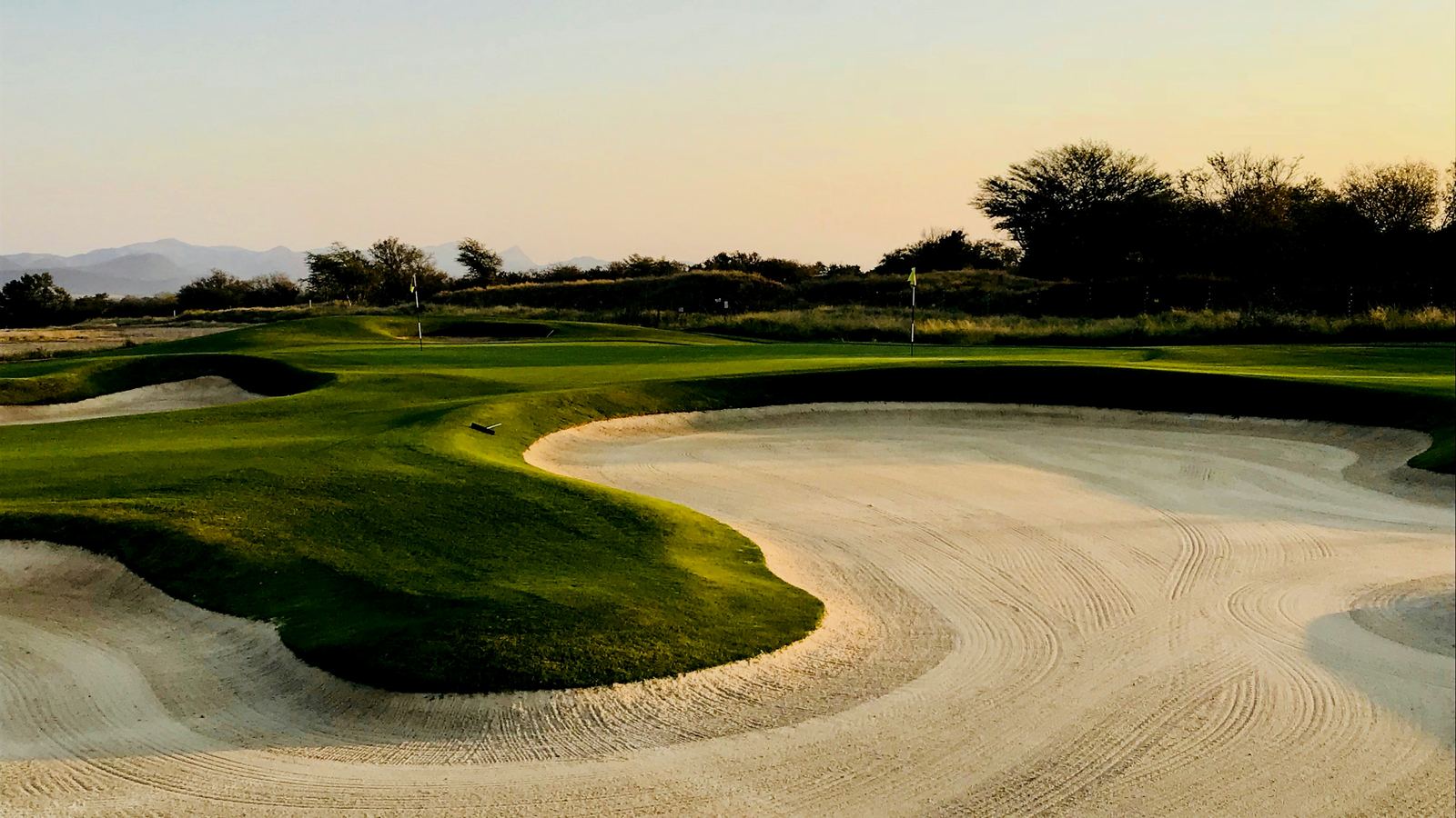 A green ringed by deep raked bunkers, late light running across the fairway and low hills on the horizon.